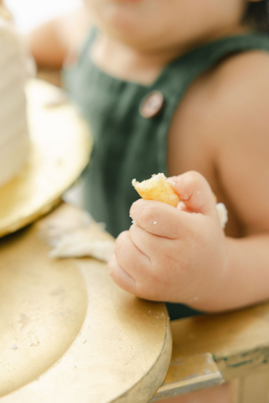 Details of an infant clutching a piece of cake while sitting in a gold high chair and green overalls from indianapolis cake shops