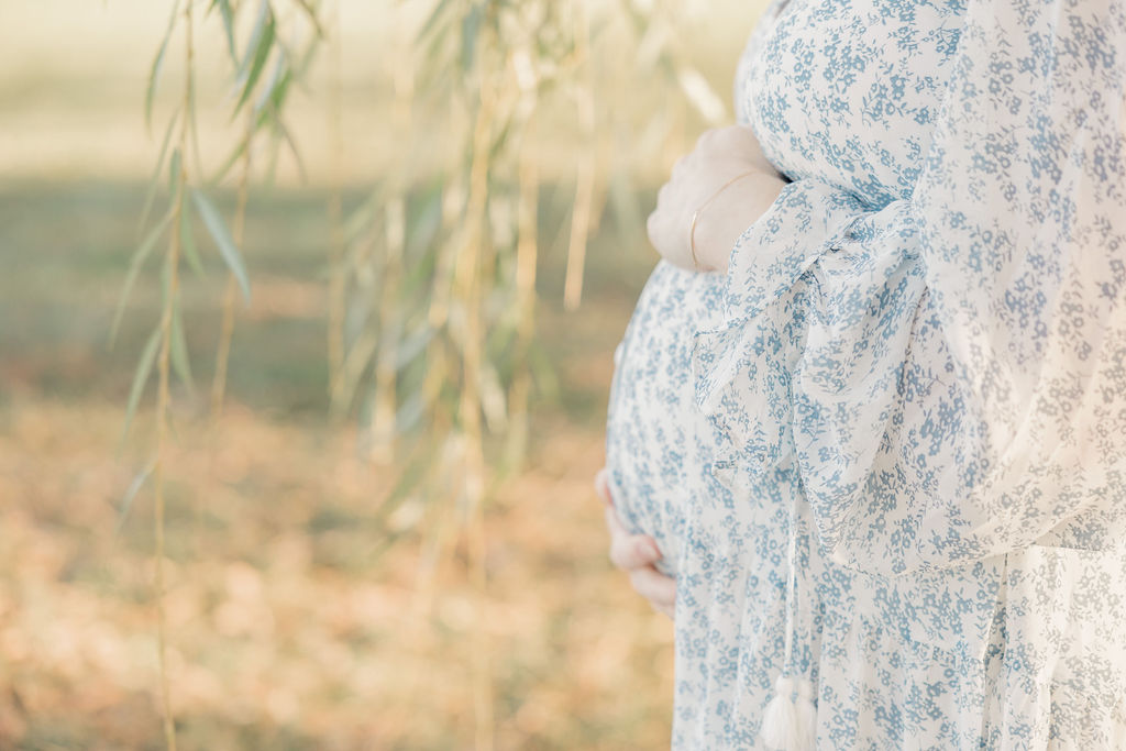 Details of a mom to be's bump while she stands under a willow tree holding her bump with both hands after visiting an indianapolis fertility clinic