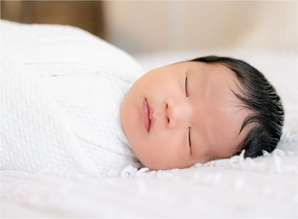 A newborn baby sleeps on a white bed and white tight swaddle