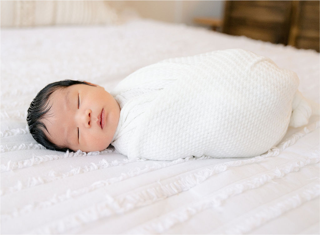 A newborn baby sleeps on a white bed in a white swaddle noblesville pediatric dentistry