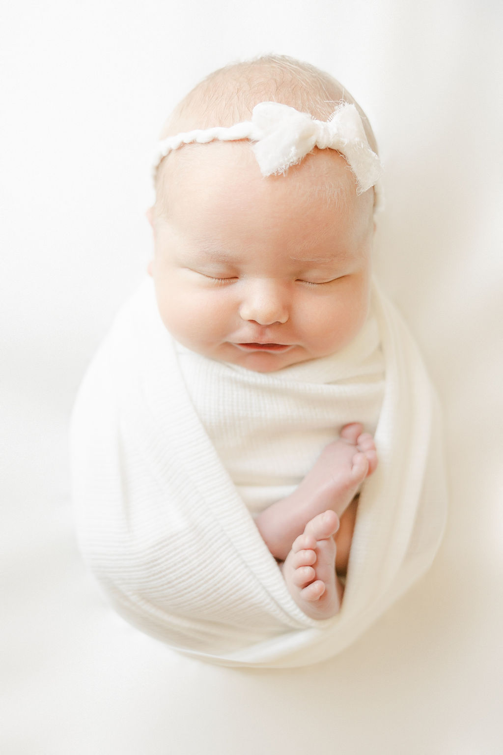 A newborn baby sleeps in a white swaddle in a studio