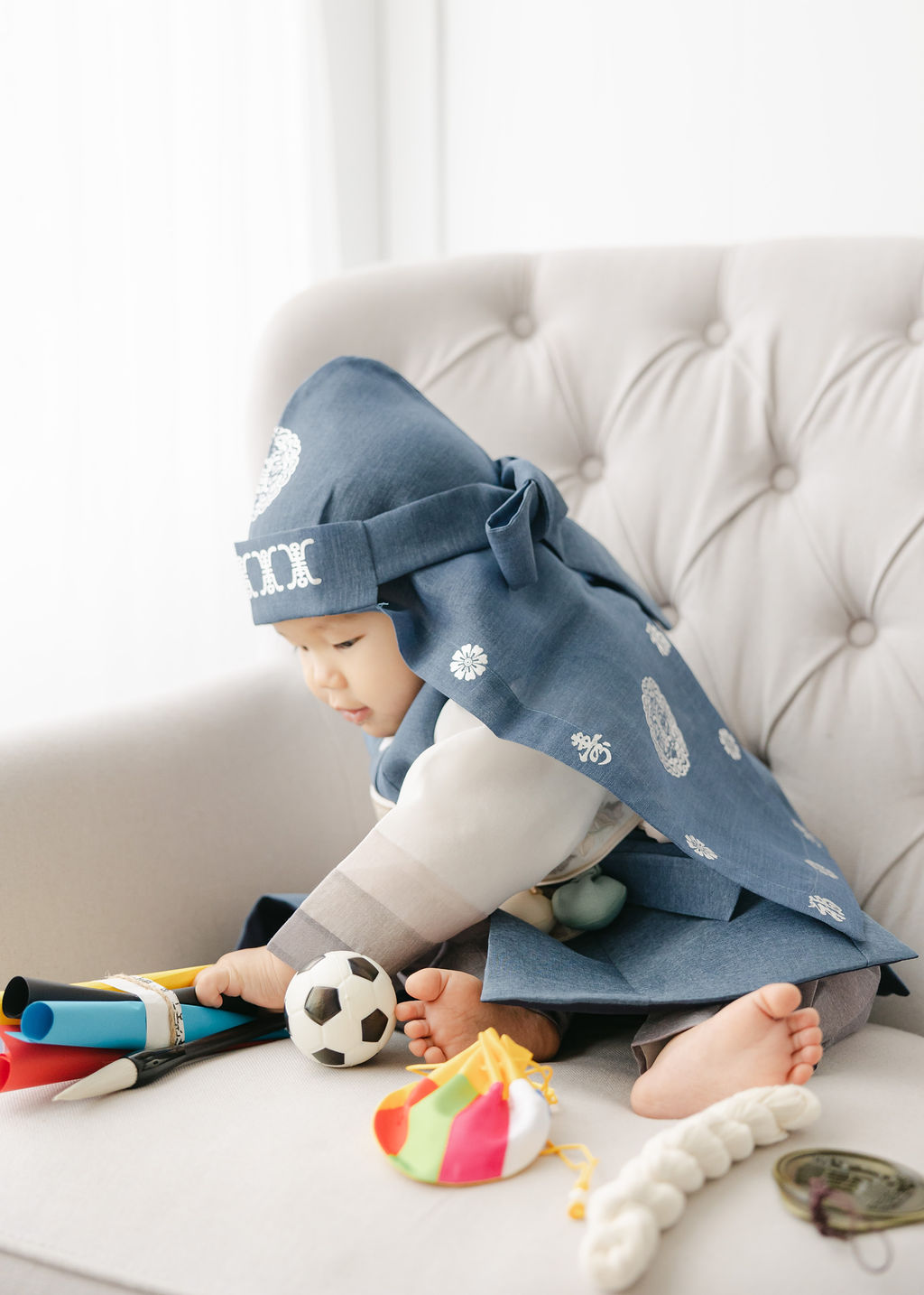 A toddler boy in a blue traditional outfit sits on a couch playing with some toys after meeting with indianapolis babysitters
