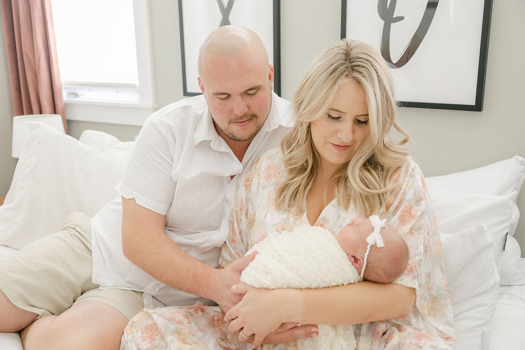 New parents sit on a white bed smiling down at their sleeping newborn baby in mom's arms