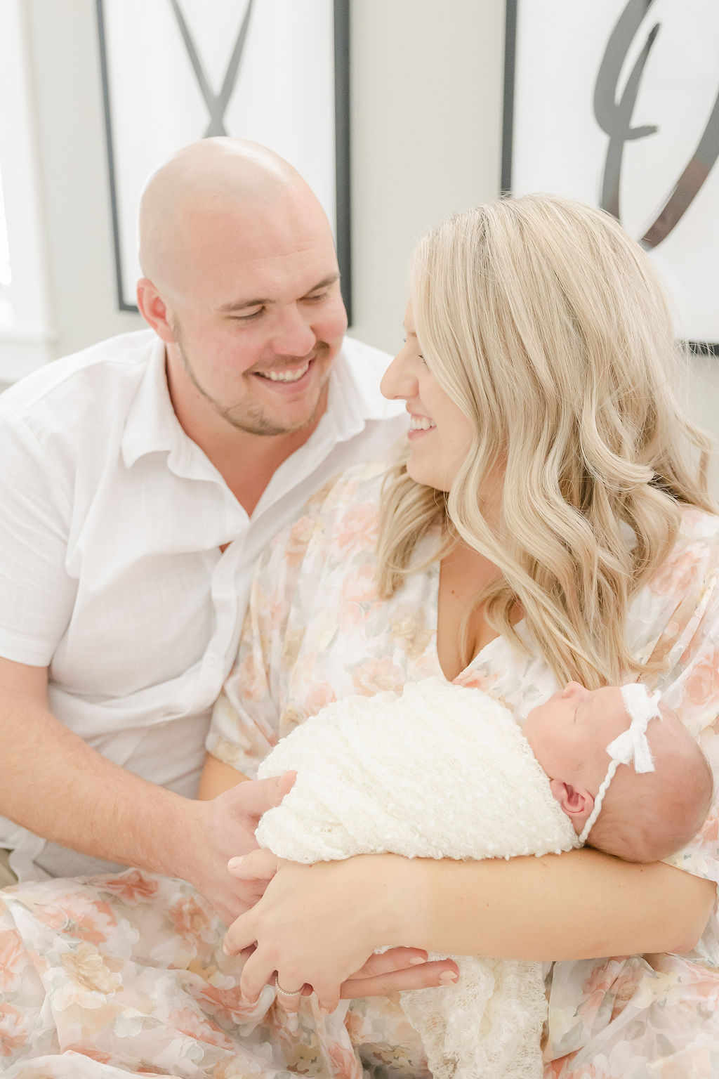 New parents smile at each other while sitting on a bed with their newborn baby sleeping in mom's arms after some indianapolis parenting classes