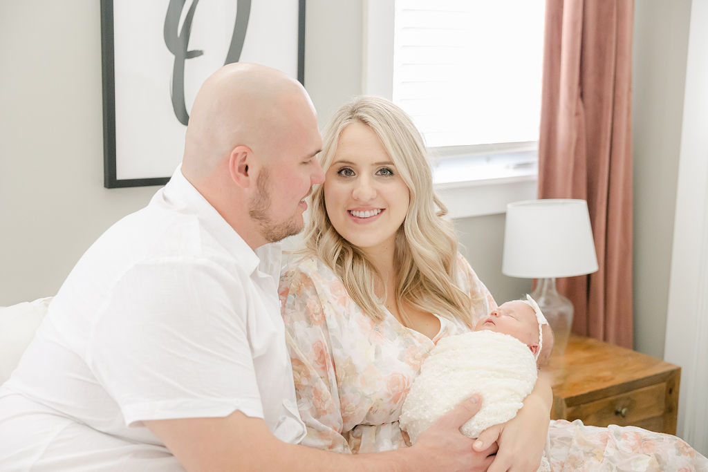 A mom and dad sit on a bed by a window holding their sleeping newborn baby after some indianapolis parenting classes