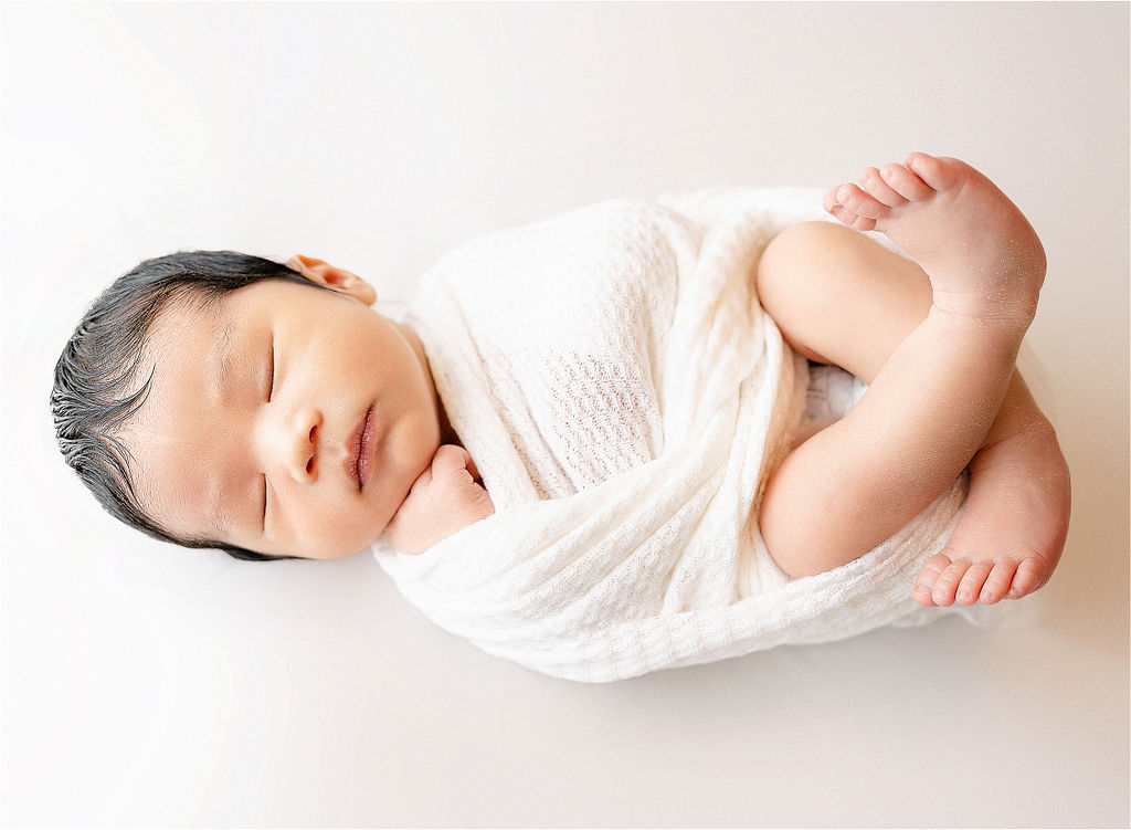 A newborn baby sleeps on a white bed wrapped in a white swaddle with legs out before visiting indianapolis pediatric dentistry