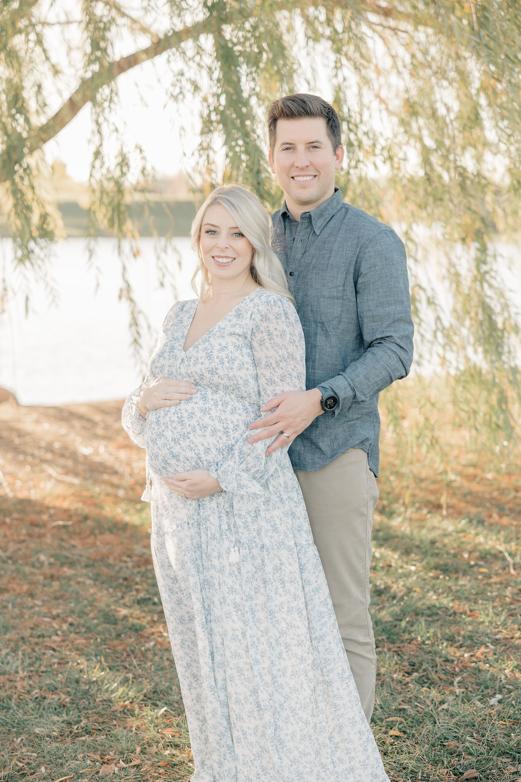 A mother to be in a blue floral print dress leans against her husband while standing under a willow tree on a lake at sunset