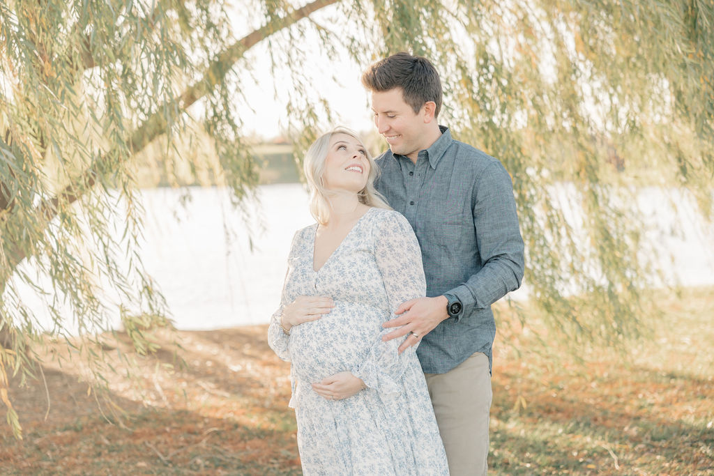 Happy expecting parents stand under a willow tree at sunset smiling at each other before some pelvic floor therapy indianapolis