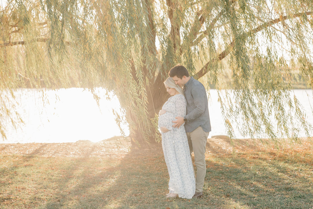 Expecting parents stand under a willow tree at sunset on a lake holding the bump before some pelvic floor therapy indianapolis