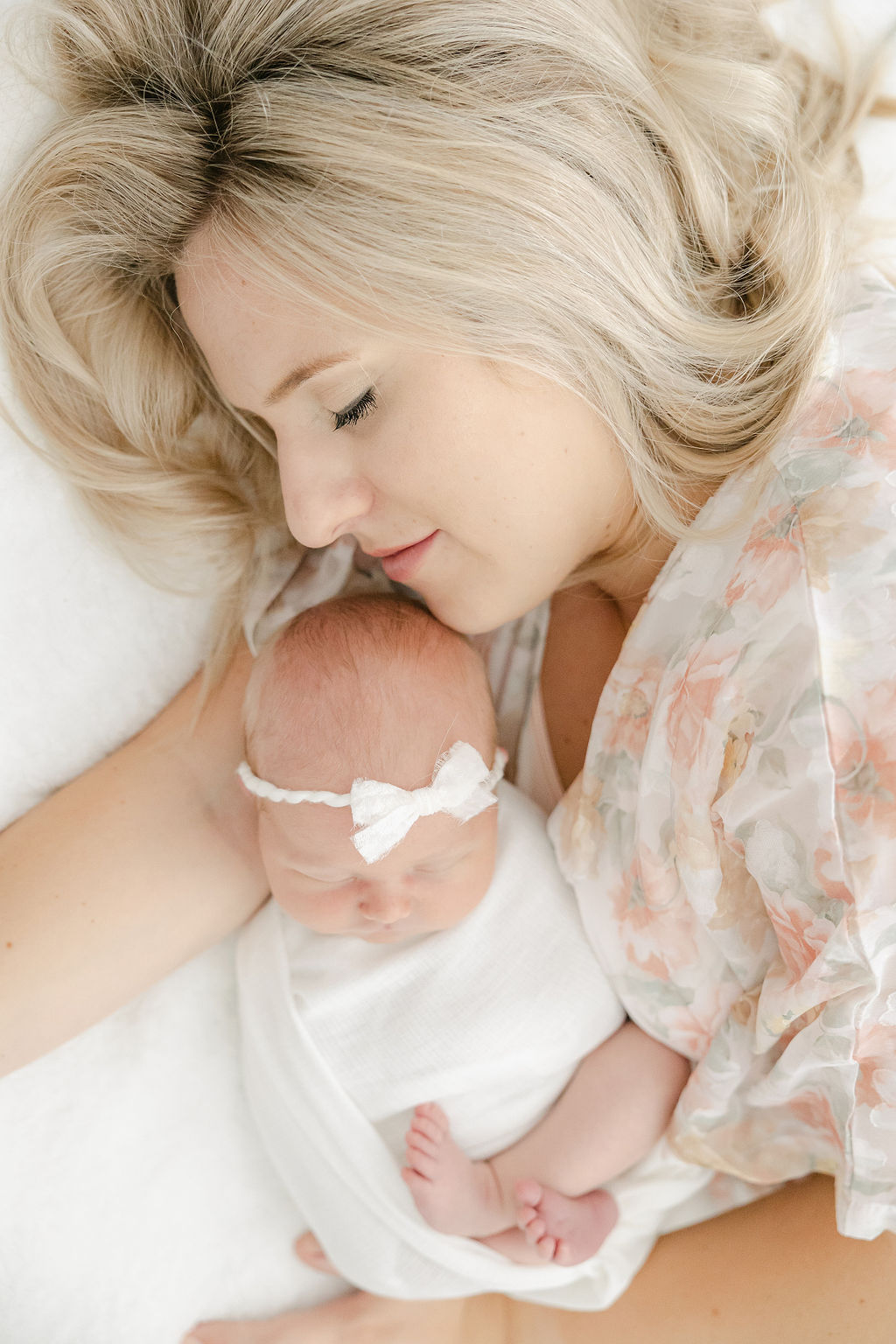 A mother in a floral dress cuddles with her sleeping newborn daughter on a white bed before visiting zionsville pediatric dentistry