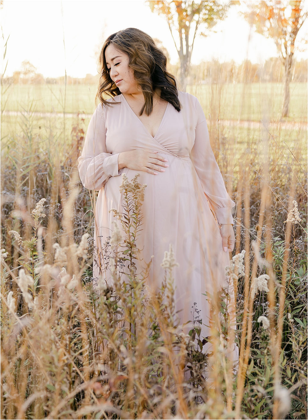 A mother to be rests a hand on her bump while walking through a field of tall wildflowers thanks to ivf indianapolis