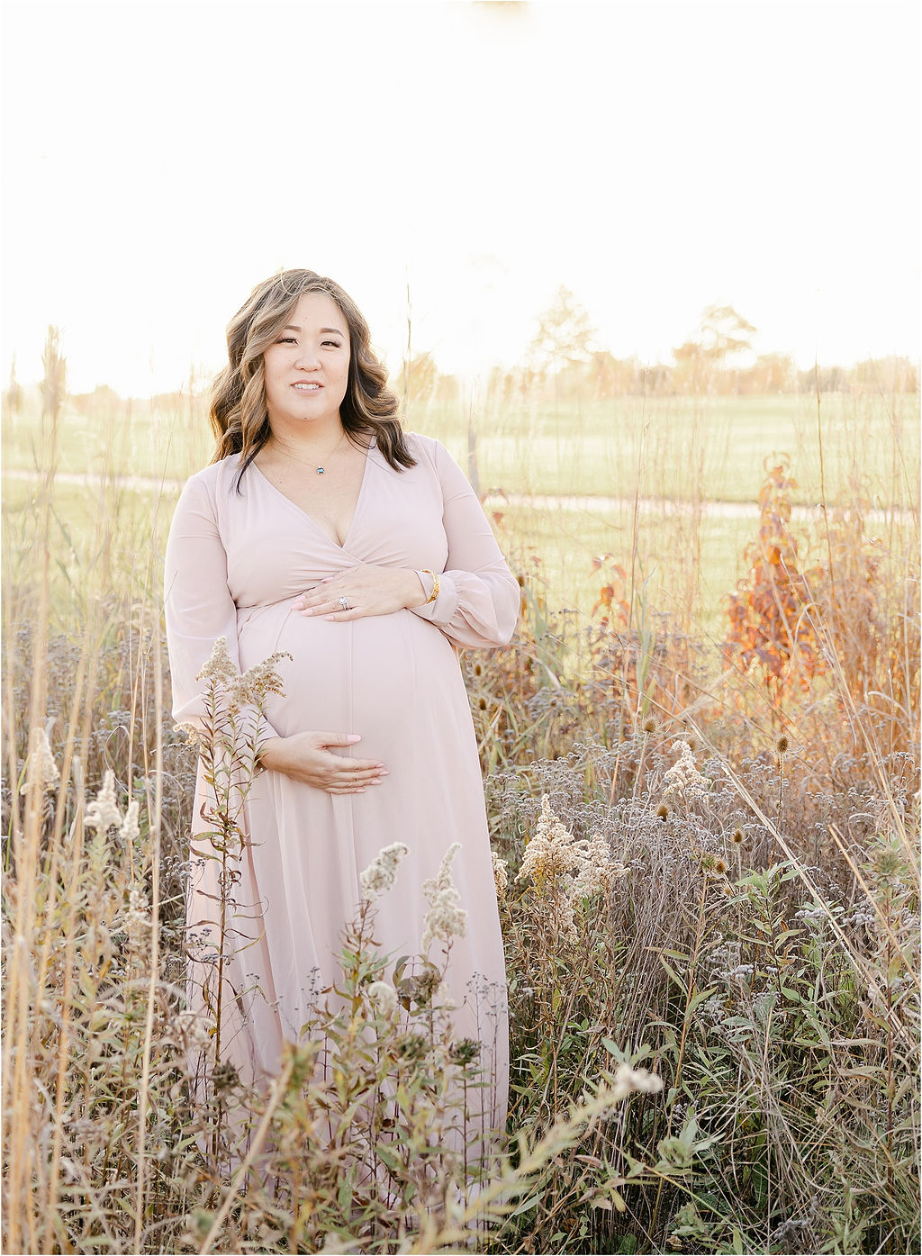 A mom to be in a pink maternity gown stands in a field of tall grasses at sunset holding her bump thanks to ivf indianapolis