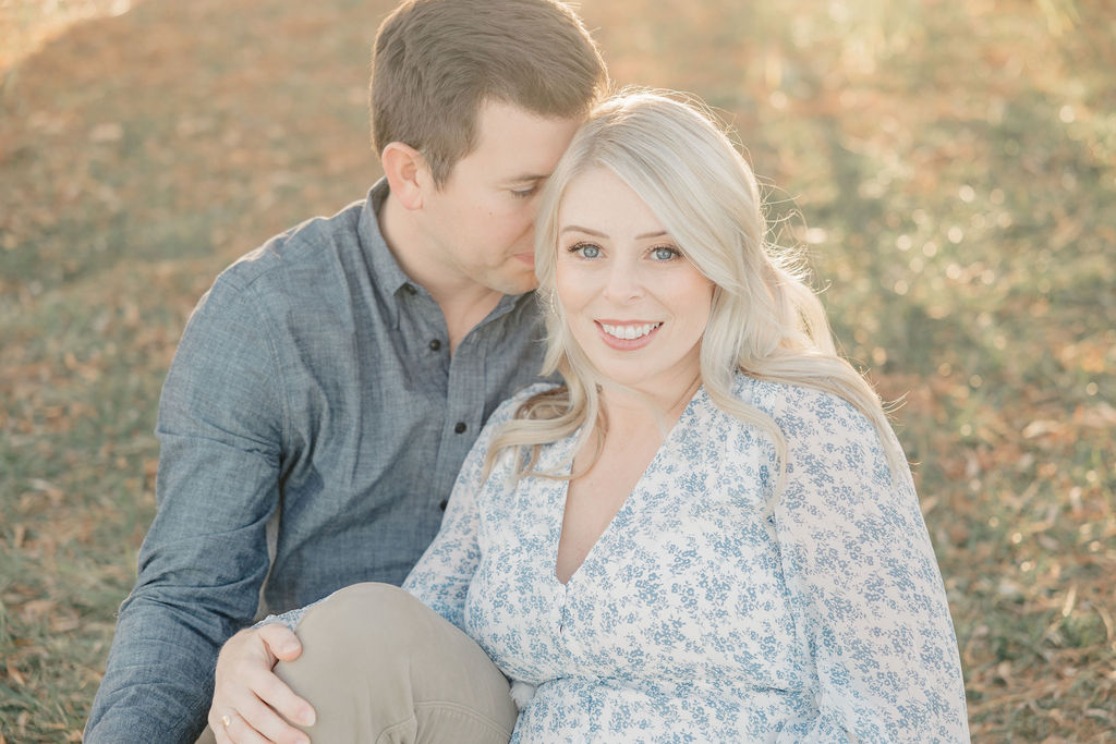Happy expecting parents sit in a park at sunset while the dad nuzzles into her blonde hair