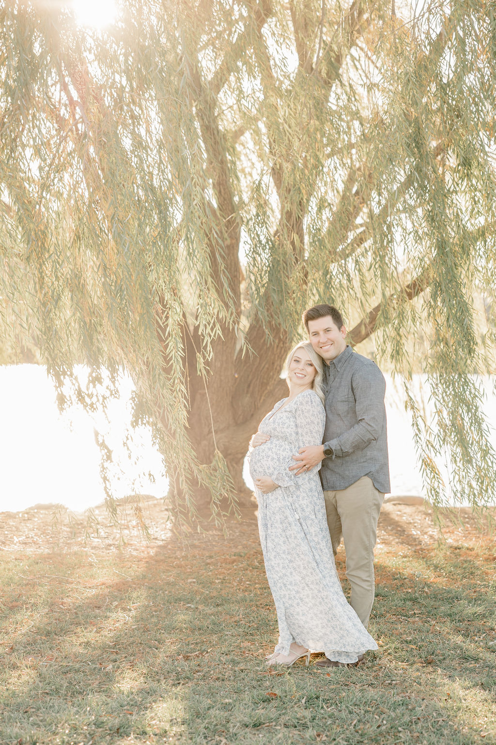 A mother to be in a blue floral print dress is hugged by by her husband from behind under a willow tree at sunset thanks to obgyn carmel indiana
