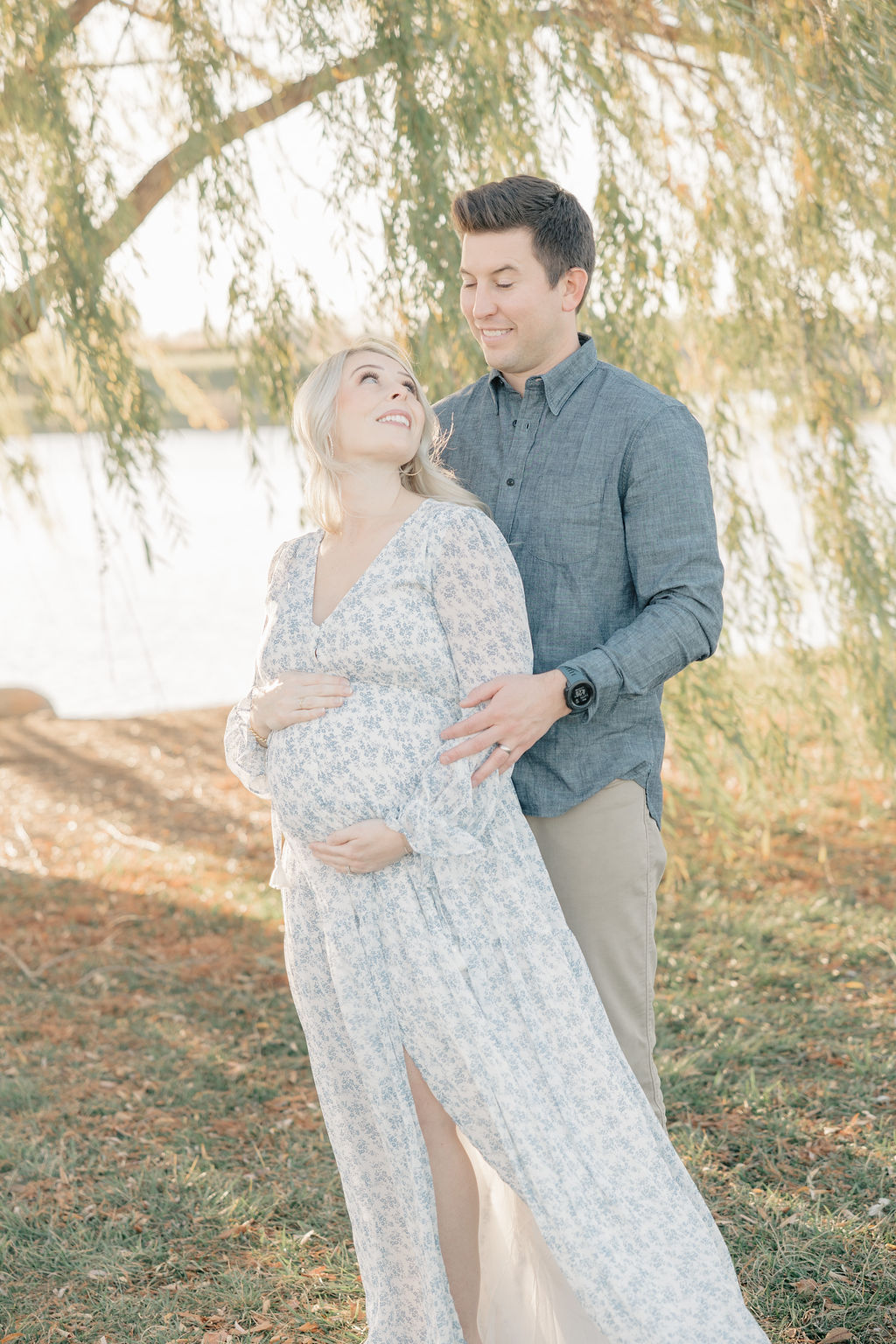 Happy expecting parents stand under a willow tree by the water smiling at each other and holding the bump thanks to obgyn carmel indiana