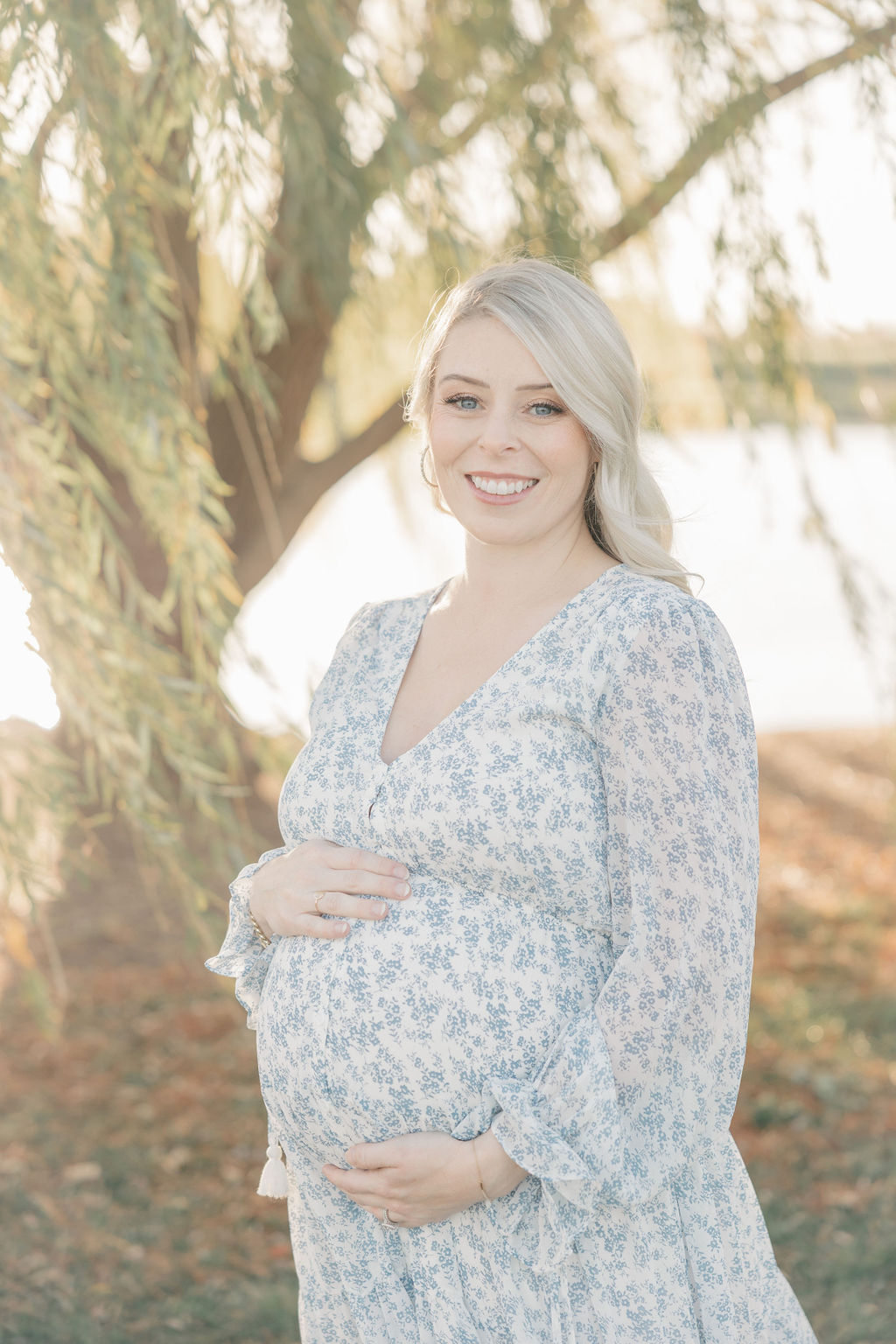 A mother to be in a blue floral dress stands smiling under a willow tree by a lake at sunset