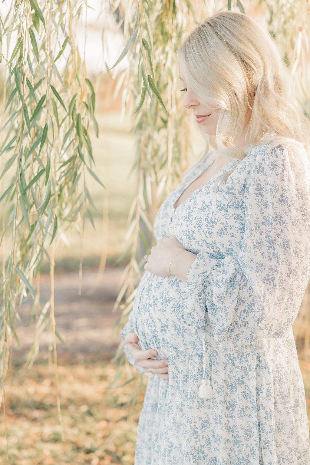 A mother to be in a blue floral dress stands under a willow tree holding her bump at sunset after visiting serene midwifery