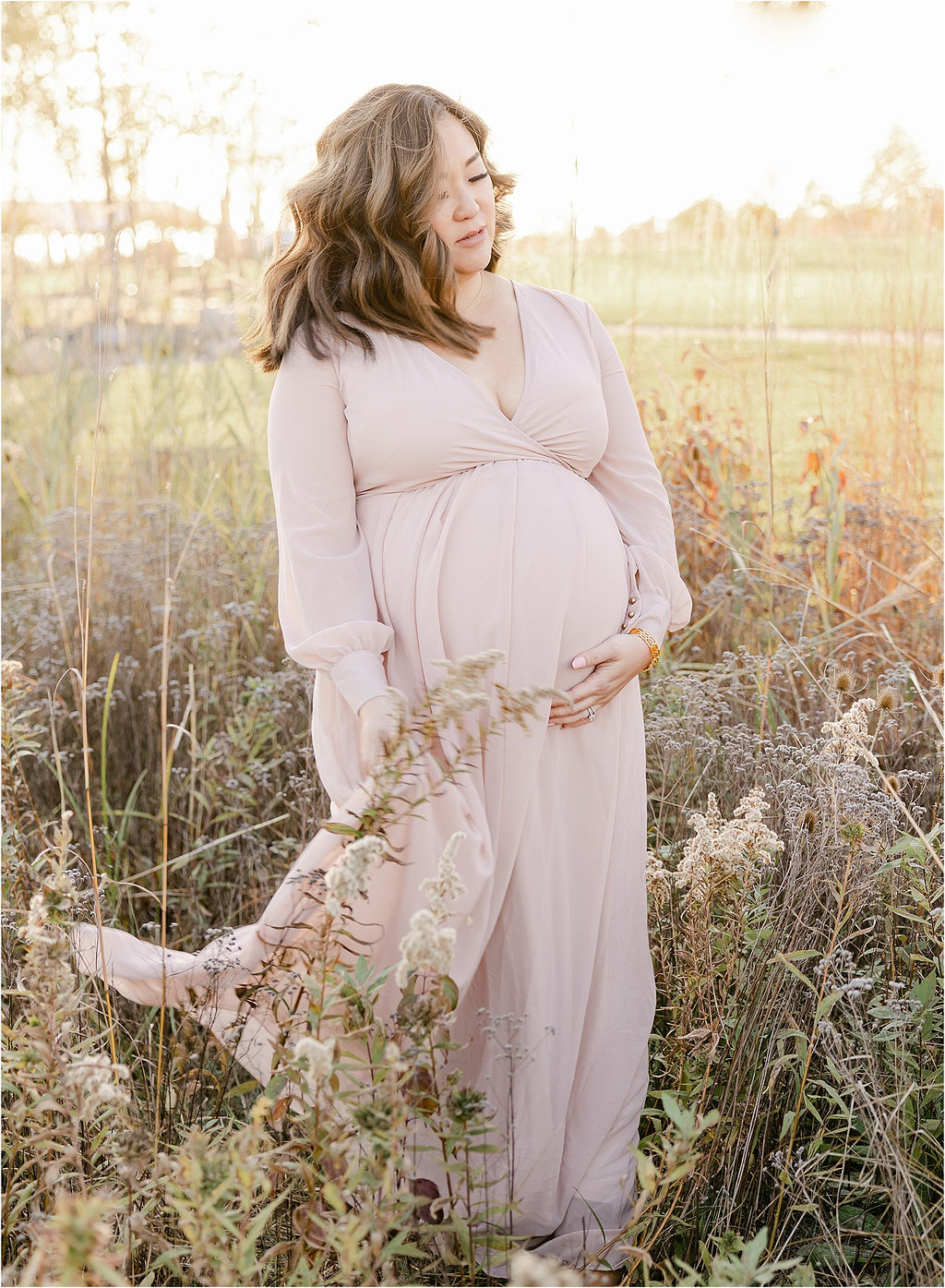 A mom to be walks through a field of tall grasses at sunset holding. her bump and train of her pink maternity gown before her water birth indianapolis