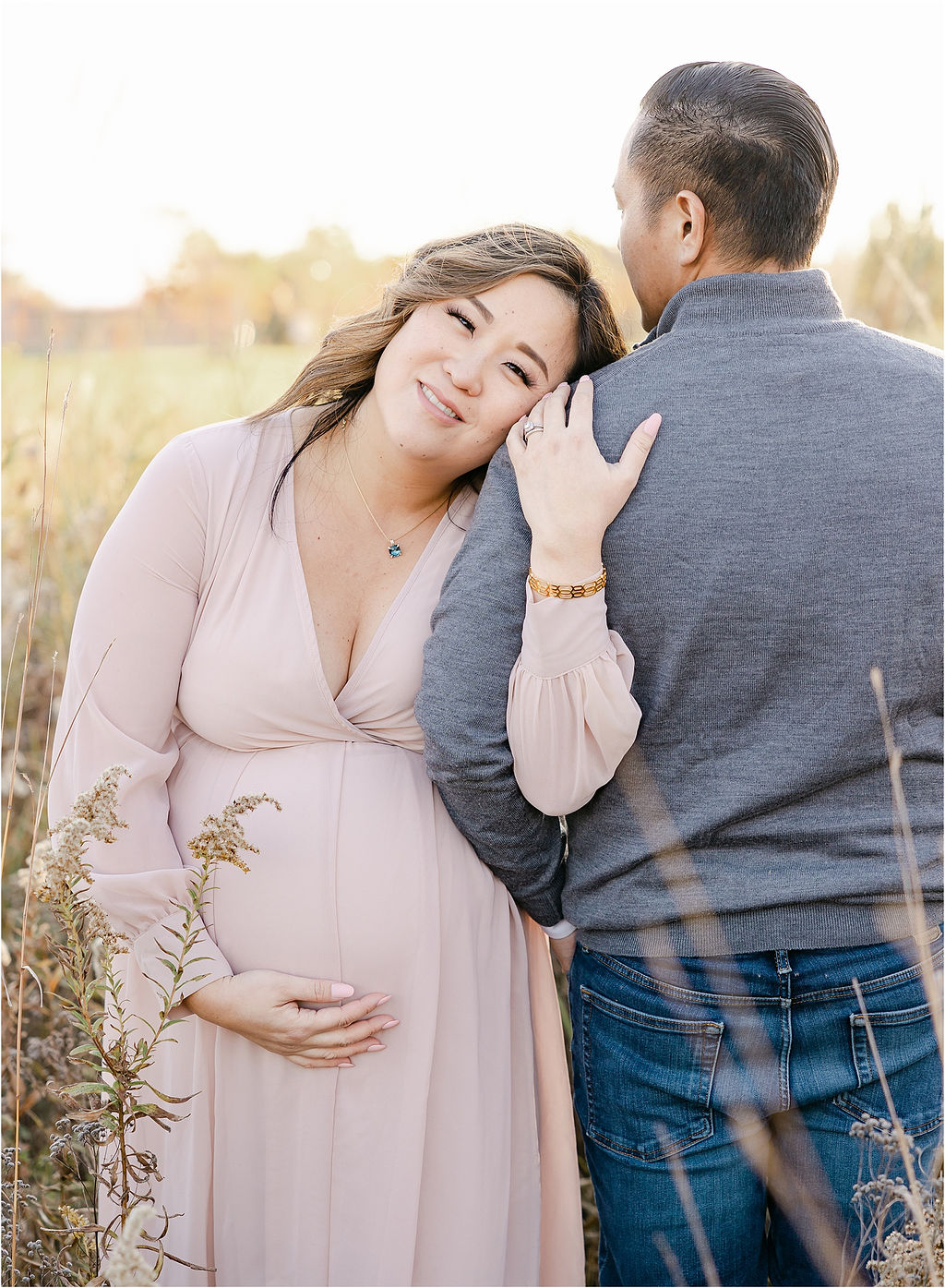 A mother to be in a pink maternity gown leans on the shoulder of her husband in a field of tall grasses at sunset before her water birth indianapolis