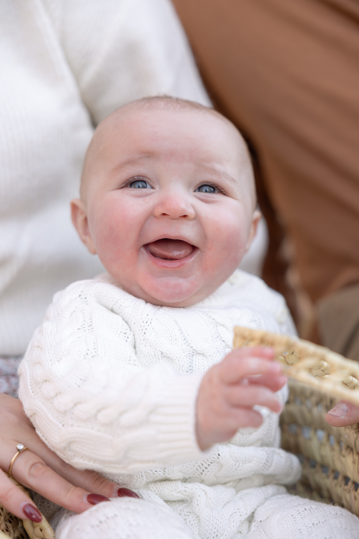 Baby in white sweater onsie smiles while sitting in basket during his session with an Indianapolis family photographer