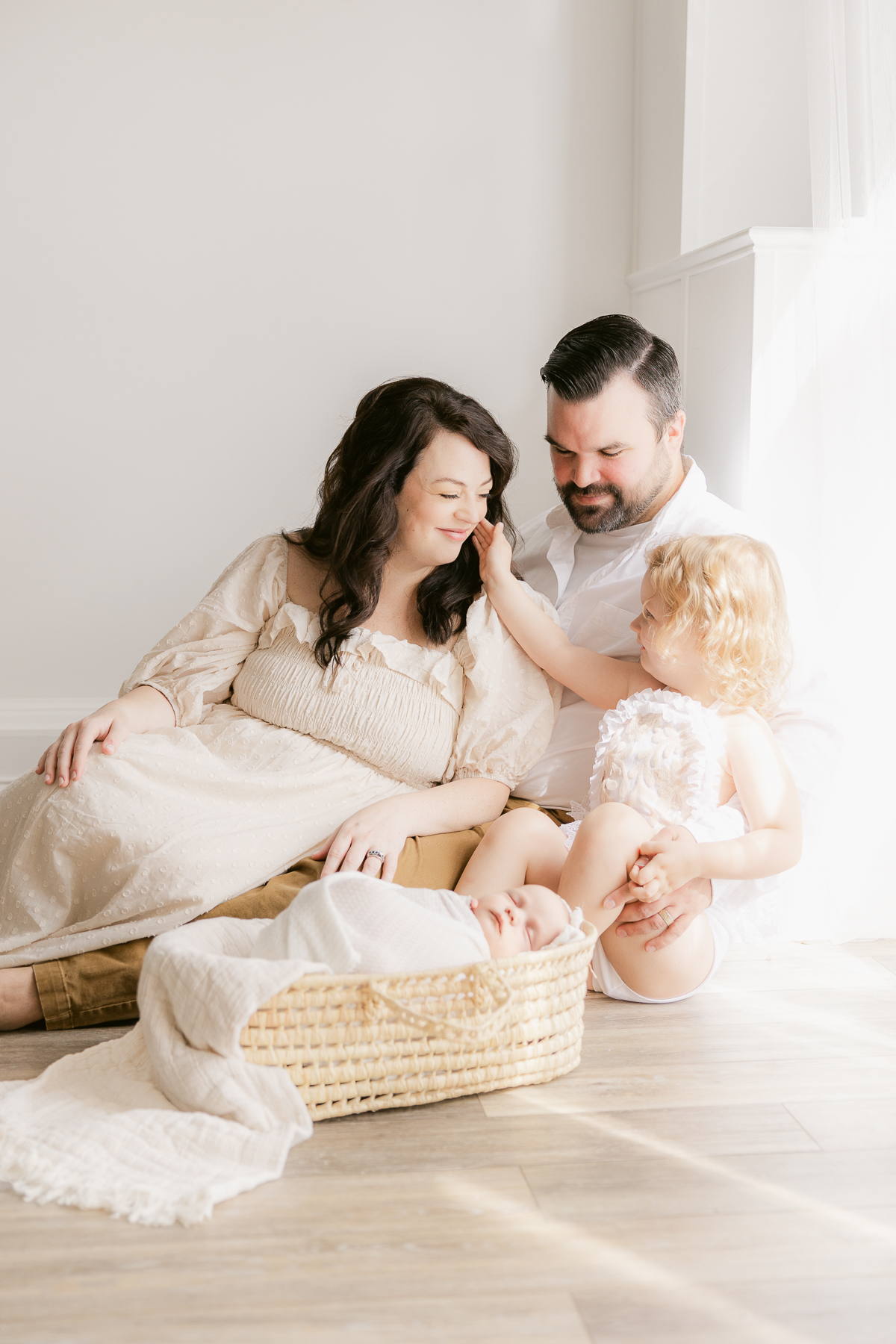 family sitting on floor in all cream clothing with newborn in a moses basket looking lovingly at eachother as toddler girl reaches up to mom, top gifts for photography loving mothers