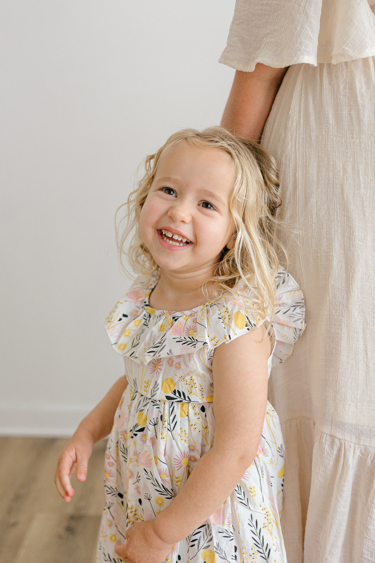 young girl in white floral dress with curly blonde hair smiles while standing close to mom in cream dress, easy photo session with kids