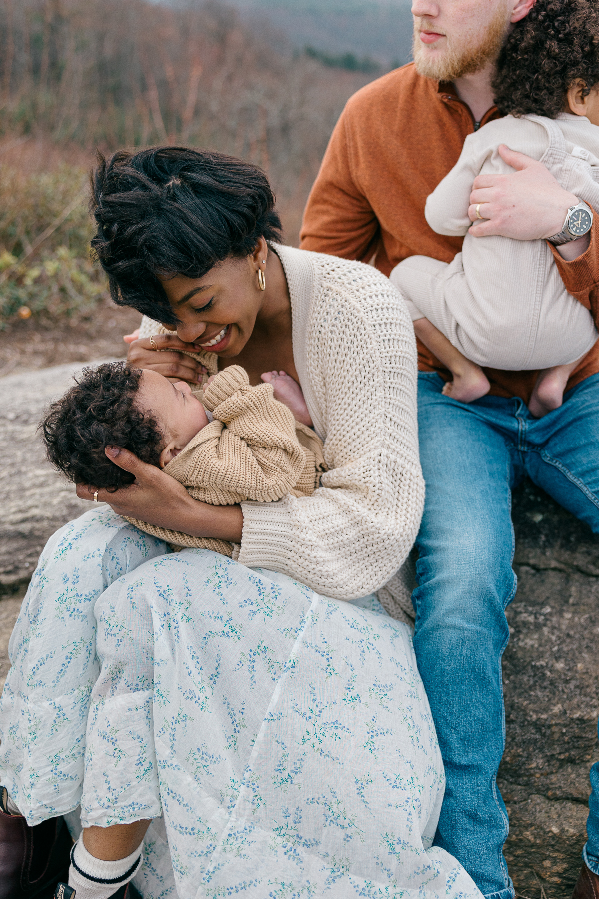 mother in blue floral dress sits holding her infant on her lap and laughing down towards him as her husband holds their toddler snuggled close to his chest behind her, easy photo session with kids