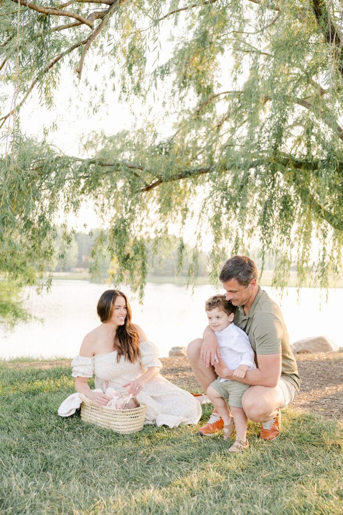 family in cream and soft green clothing sits on the grass under a willow tree in front of a pond with their newborn baby in a moses basket near mom while dad wraps his arms around their toddler son, family photography in Carmel