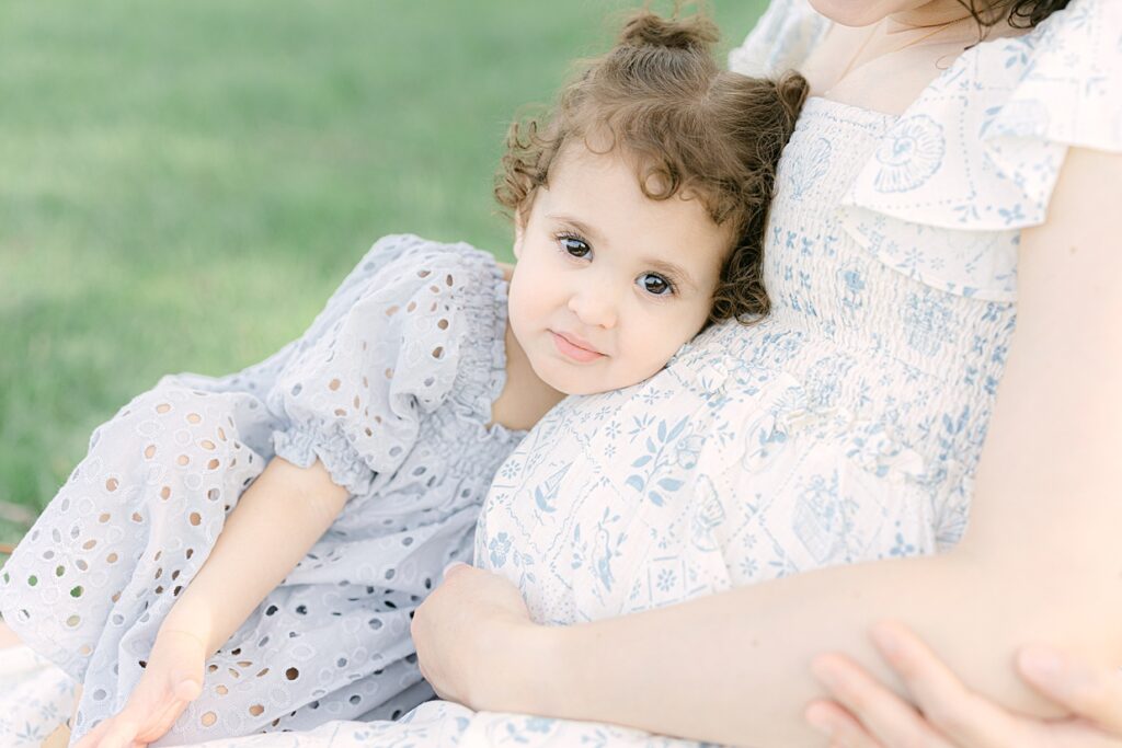 toddler girl lays her head on her pregnant mother's baby bump and looks into the camera in a light blue textured dress showing a great example of what to wear for spring family pictures