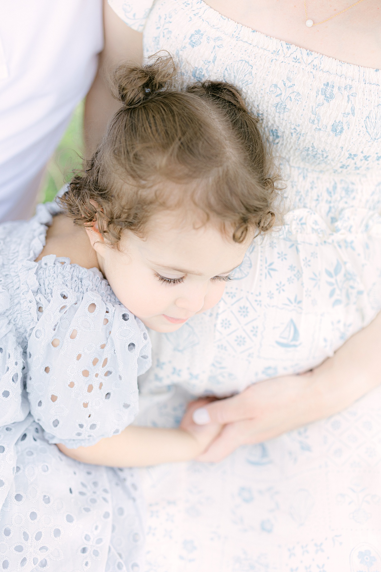 toddler girl lays her head on her pregnant mother's baby bump while holding her hand in a light blue textured dress showing a great example of what to wear for spring family photos