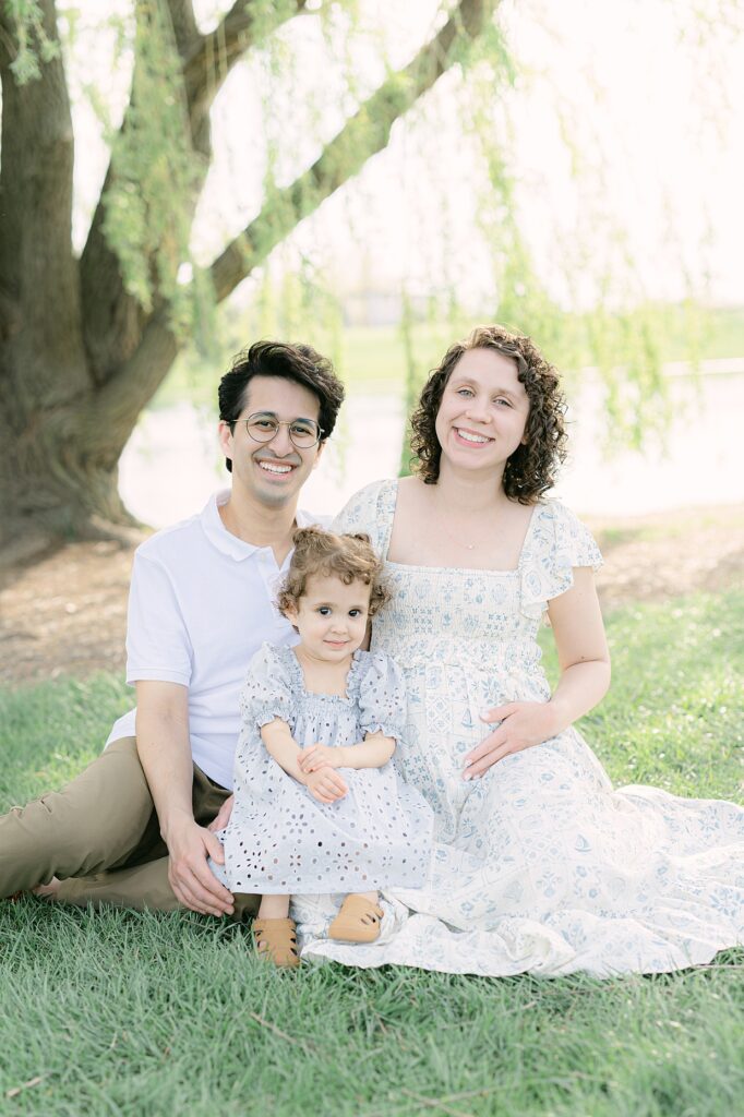 a young family sits in the grass under a willow tree wearing creams and light blues with mom in a floral pattern and her toddler in a textured dress while dad wears neutrals for their spring family photos