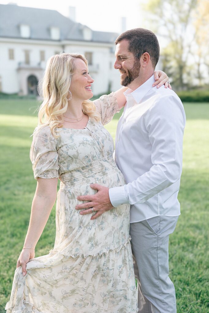 A pregnant woman in a floral sage green dress looks at her husband dressed in white and grey during their Indianapolis spring family pictures