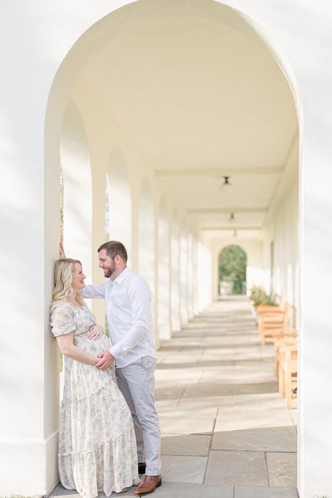 Pregnant mother in a sage green floral dress leans against a cream pillar while her husband holds her hand on her baby bump wearing a white button up and light grey slacks to show what to wear for spring family photos