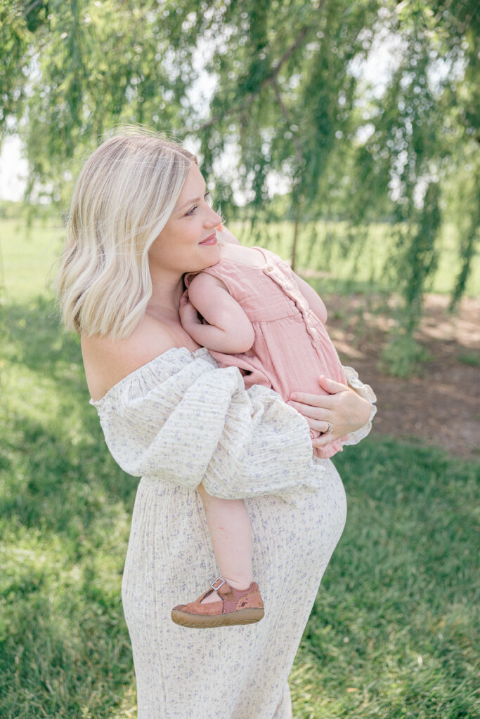 Pregnant mother in a cream dress with a purple floral pattern holds her toddler dressed in pink during their spring family photos