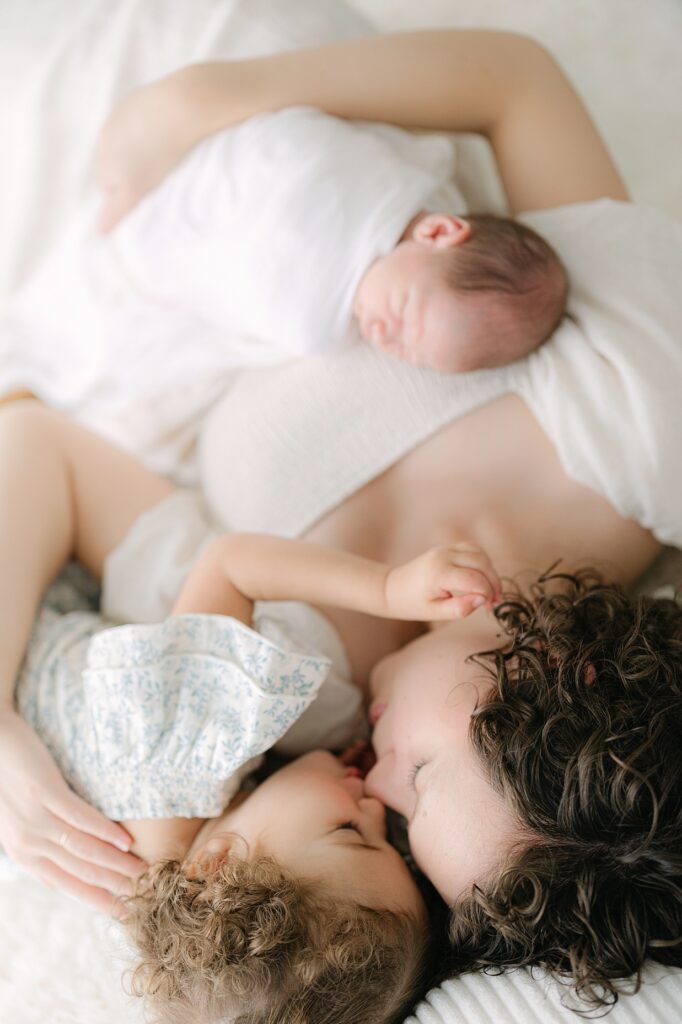 a mother in a cream dress holds her newborn swaddled in a white blanket while laying next to her toddler daughter nose to nose during a studio newborn session