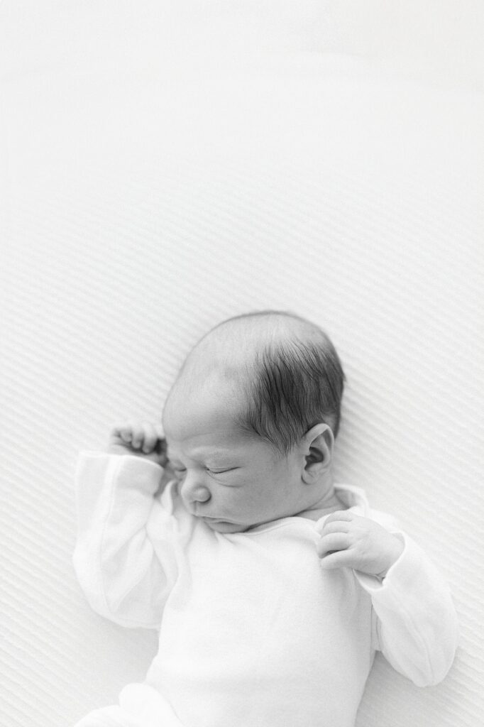 Black and white photo of a newborn baby in a white onsie laying on a white bed sleeping with his hand curled by his face during a studio newborn session