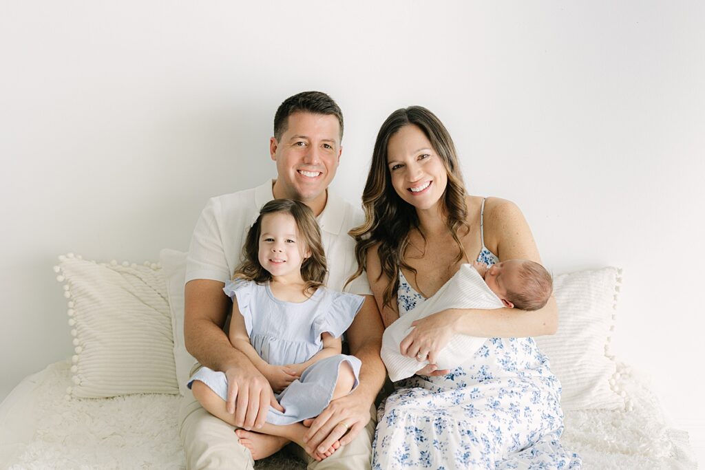 A family of 4 sits on a bed smiling during a newborn session with dad holding his toddler daughter and mom holding their newborn baby boy all in white, cream, and light blues