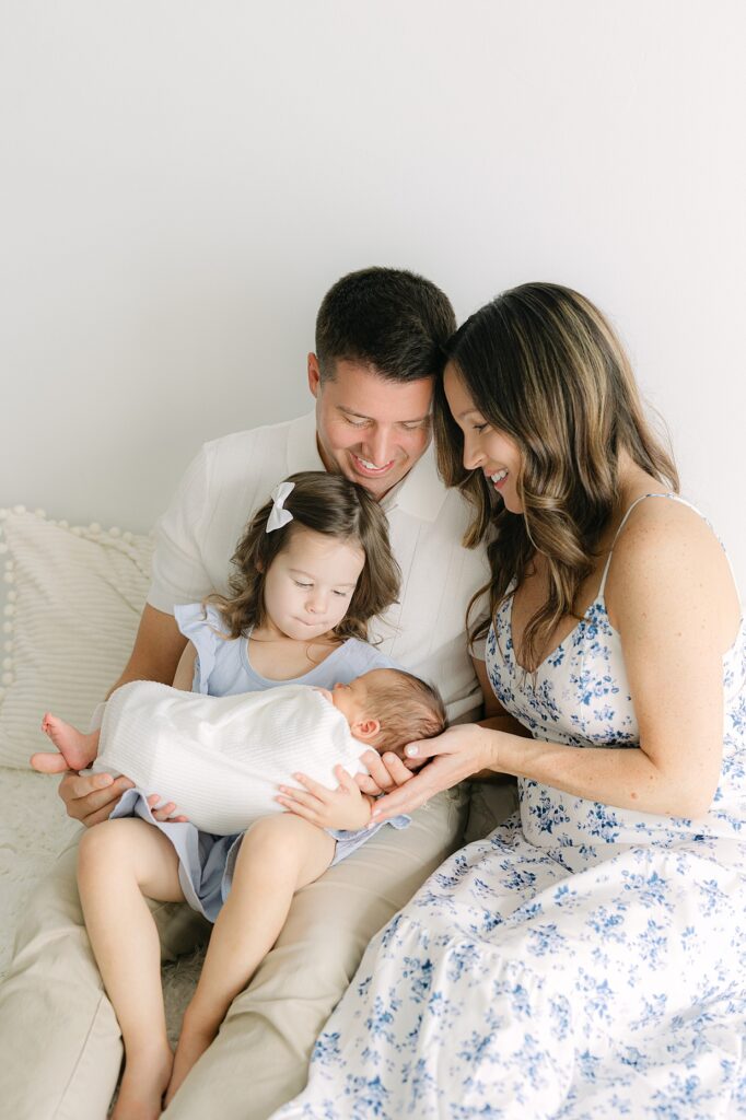 A family of four sits on a bed, mom and dad leaning into each other, their toddler daughter on dad's lap as she holds her newborn baby brother swaddled in white for their Indianapolis newborn session