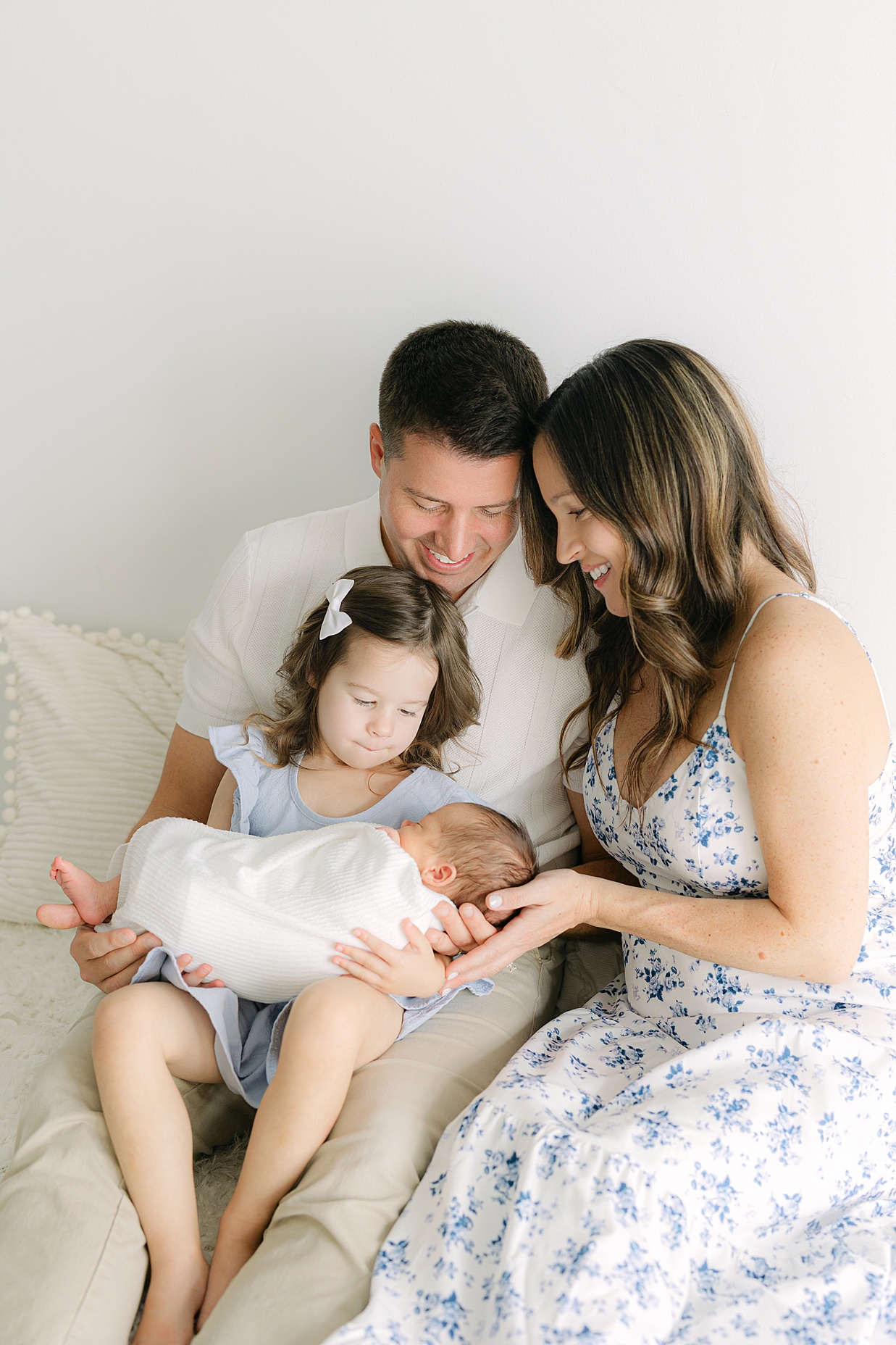 A family of four sits on a bed, mom and dad leaning into each other, their toddler daughter on dad's lap as she holds her newborn baby brother swaddled in white for Indianapolis family pictures