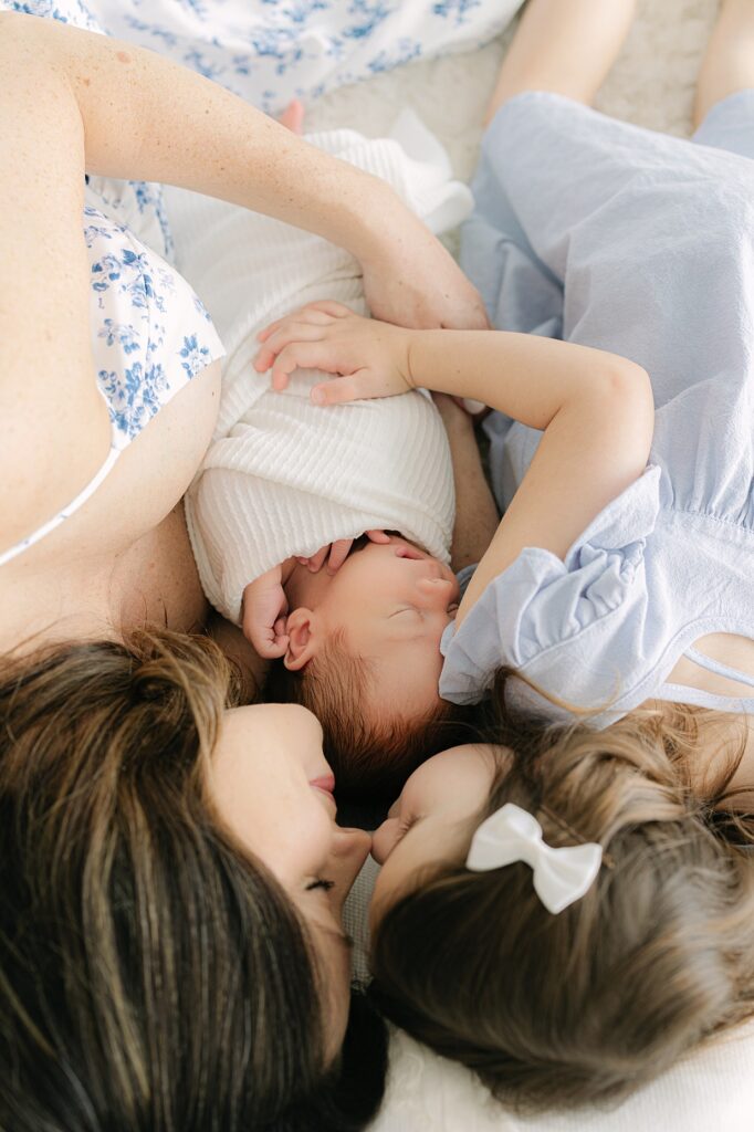 A mother in a white dress with blue flowers lays with her toddler daughter in a blue dress snuggling noses with her newborn son sleeping between them during their Indianapolis newborn session