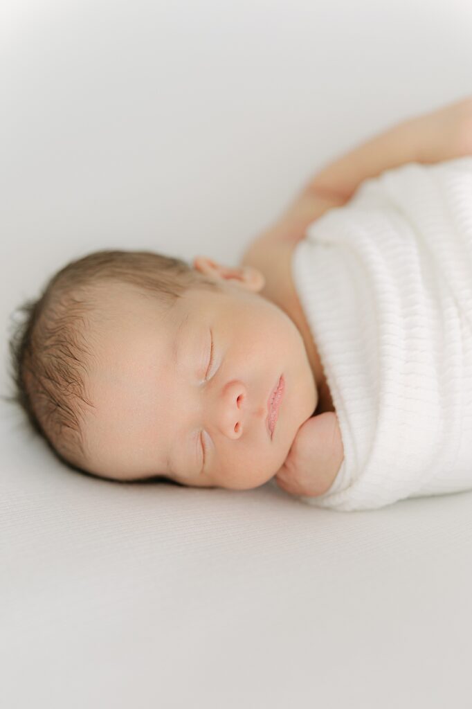baby swaddled in white blanket sleeps with his fist under his chin during his Indianapolis newborn session