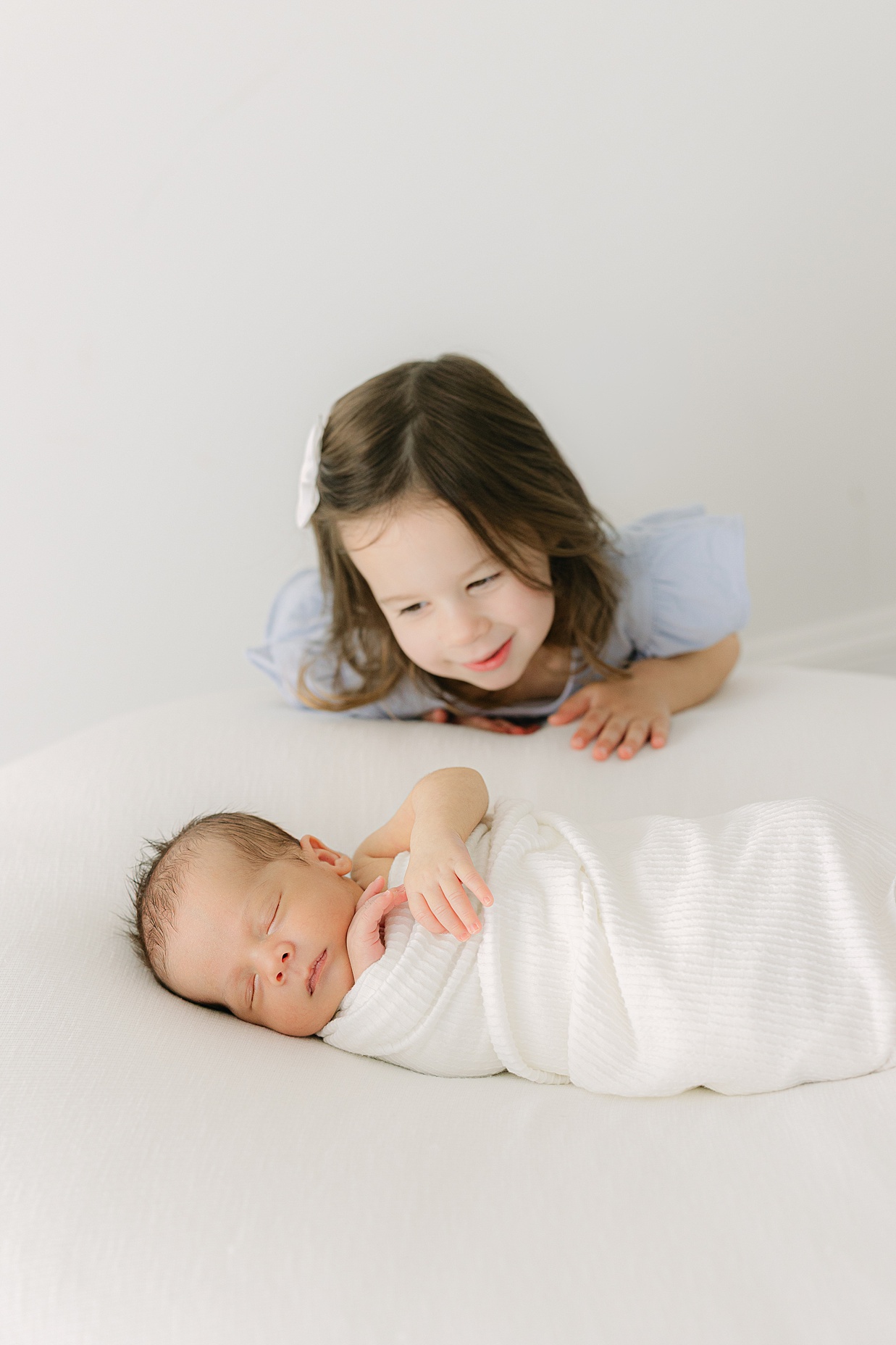 Toddler girl looks lovingly at her newborn brother swaddled in white laying on a white bed during a studio newborn session