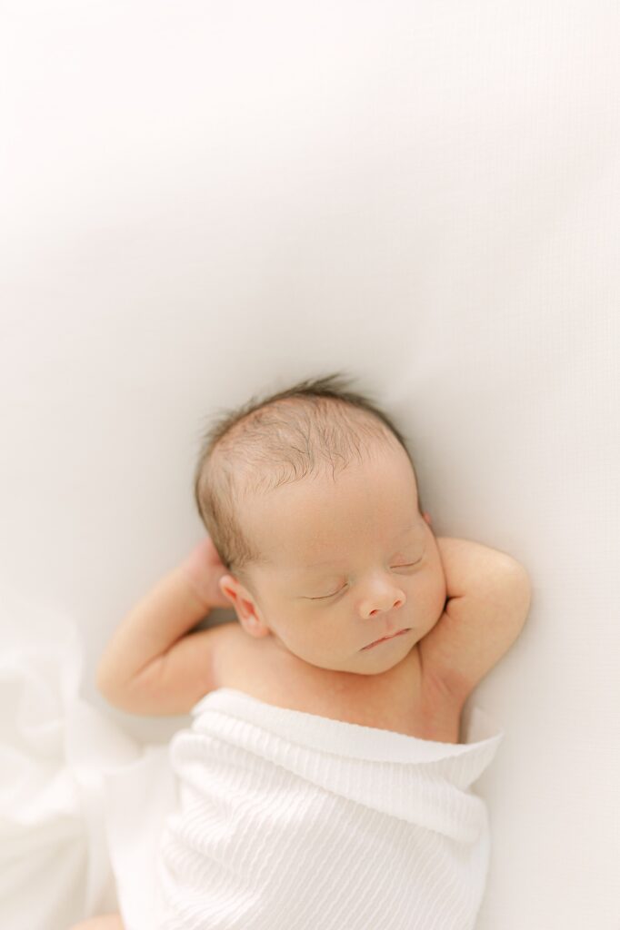 Baby sleeping on a white bed wrapped in a white blanket with his arms behind his head at his Indianapolis newborn session