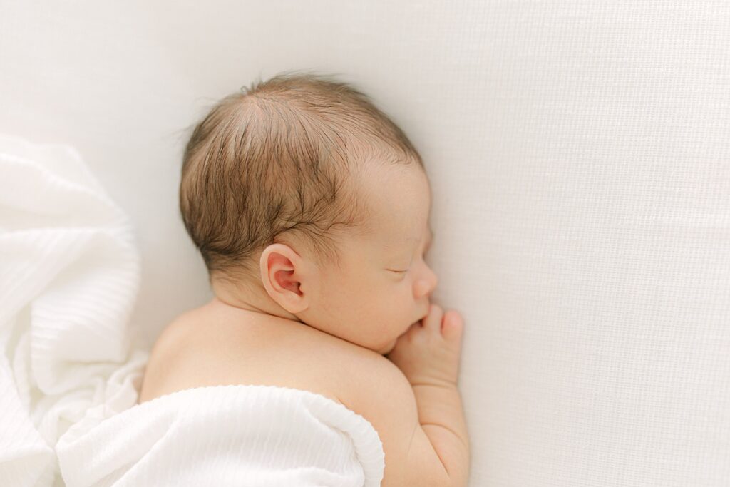 Newborn boy laying on a white bed on his stomach with his hand by his mouth covered in a white blanket during a studio newborn session