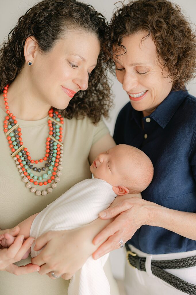 A mother in a light green dress holds her newborn baby swaddled in white while smiling at him with his grandma snuggling in and smiling at him during their Indianapolis newborn session