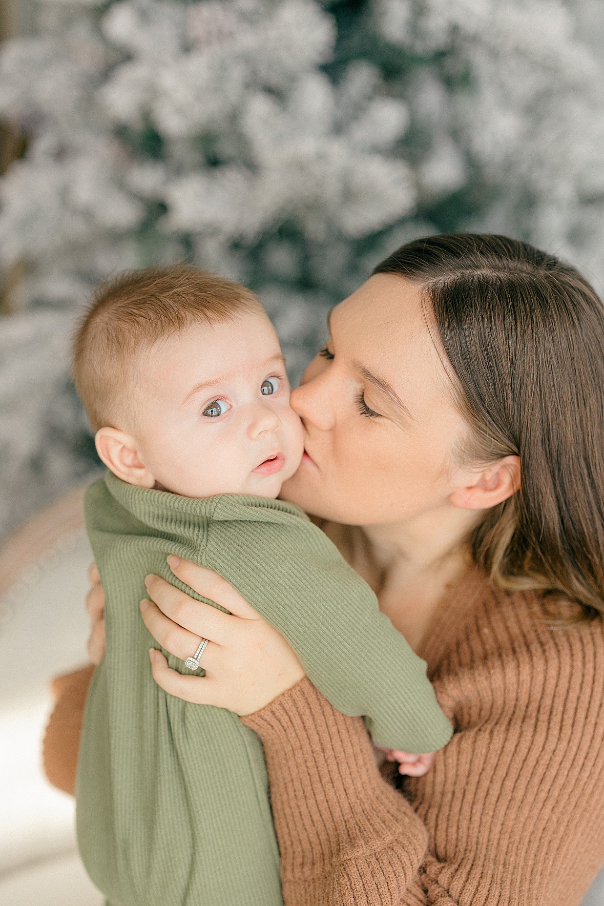 Ideas for what to Wear for Christmas Family Photos mother in brown sweater dress kisses her baby boy dressed in a green sweater onsie in front of a flocked tree