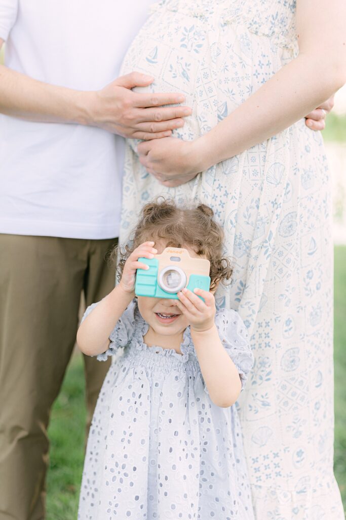 Toddler girl in blue dress stands in front of her parents holding a wooden camera over her face during Indianapolis family pictures