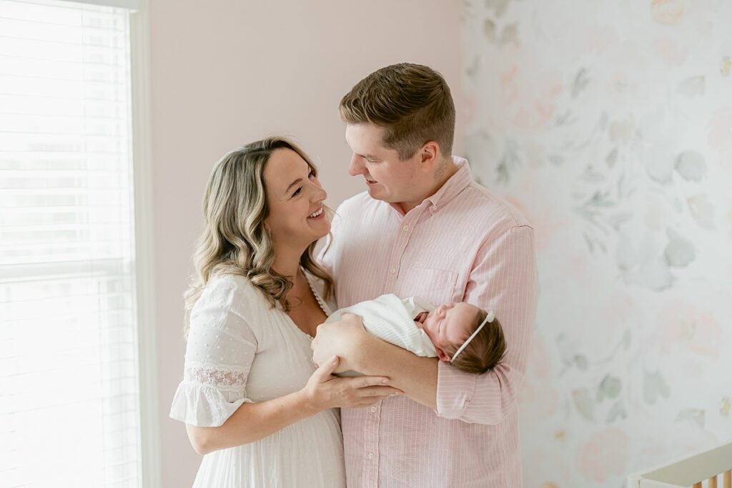 Mother and father smile at each other while dad holds his newborn baby daughter in their nursery with pink and sage florals, Indianapolis newborn session
