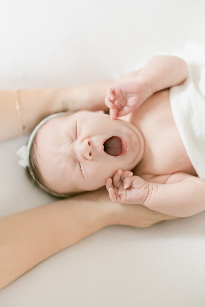 Newborn swaddled in white with a white bow in her hair yawns with her hands by her face during a newborn session